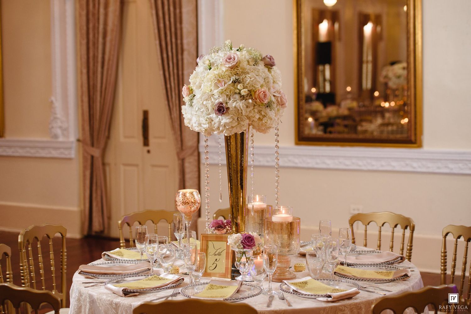 Elegant wedding reception table with tall floral centerpiece, gold accents, crystal glasses, and hanging pearl strands in a grand ballroom.