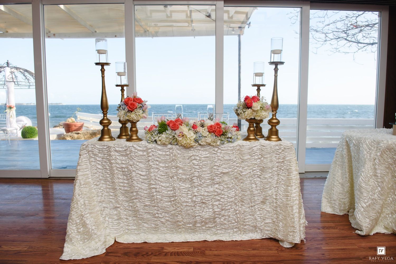 Elegant wedding sweetheart table with gold candelabras, coral floral arrangements, and white textured linen overlooking the ocean.