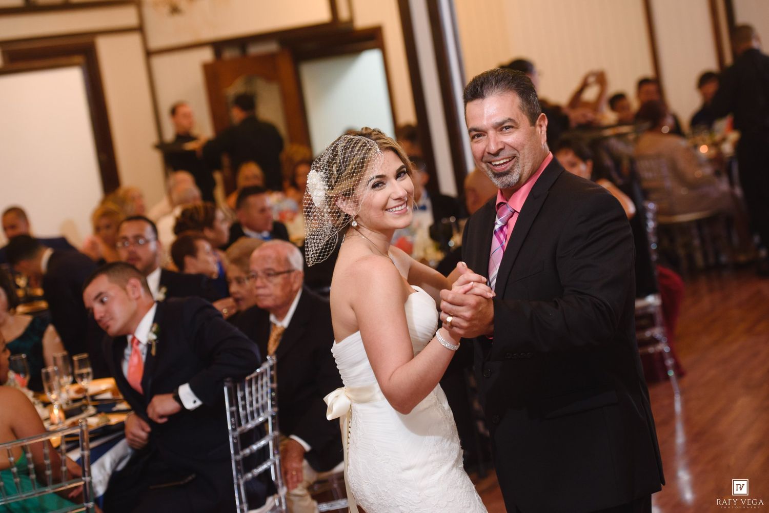 Bride and groom share a romantic first dance at their wedding reception while guests celebrate in the background.