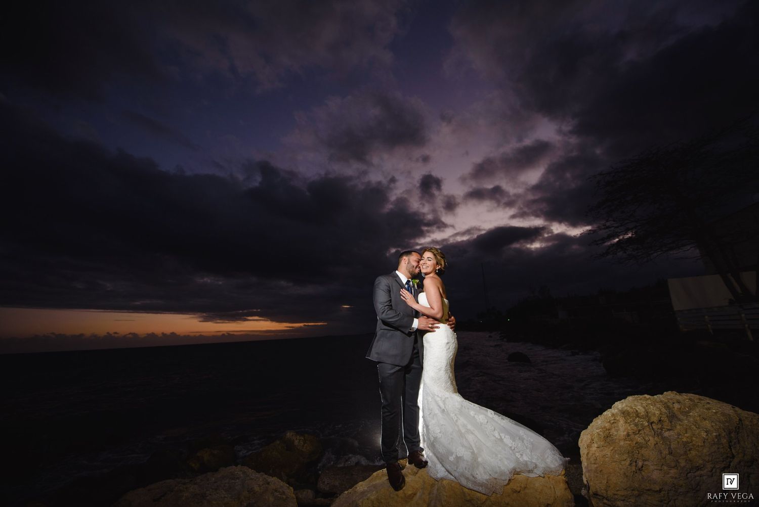 Bride and groom embrace on rocky shore at dramatic sunset with stormy purple sky backdrop.