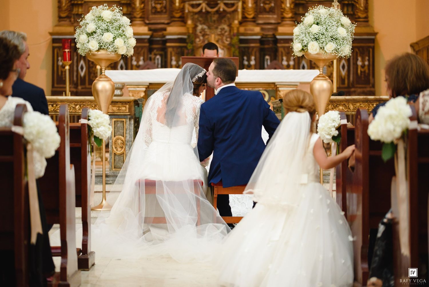 Bride and groom share a kiss at the altar during a traditional church wedding ceremony with white floral arrangements.