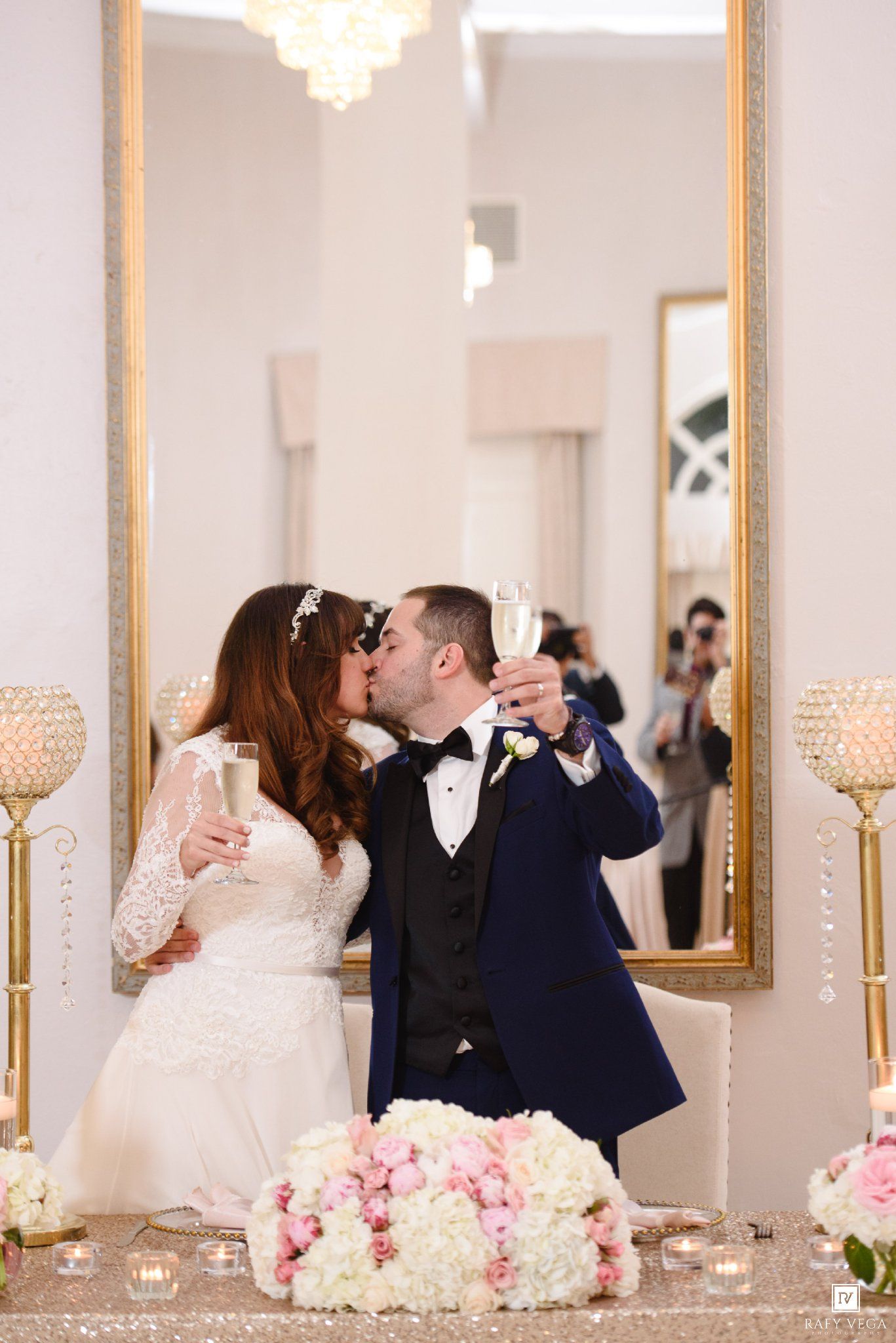 Bride and groom kissing while holding champagne glasses in front of a gold mirror at their elegant wedding reception.