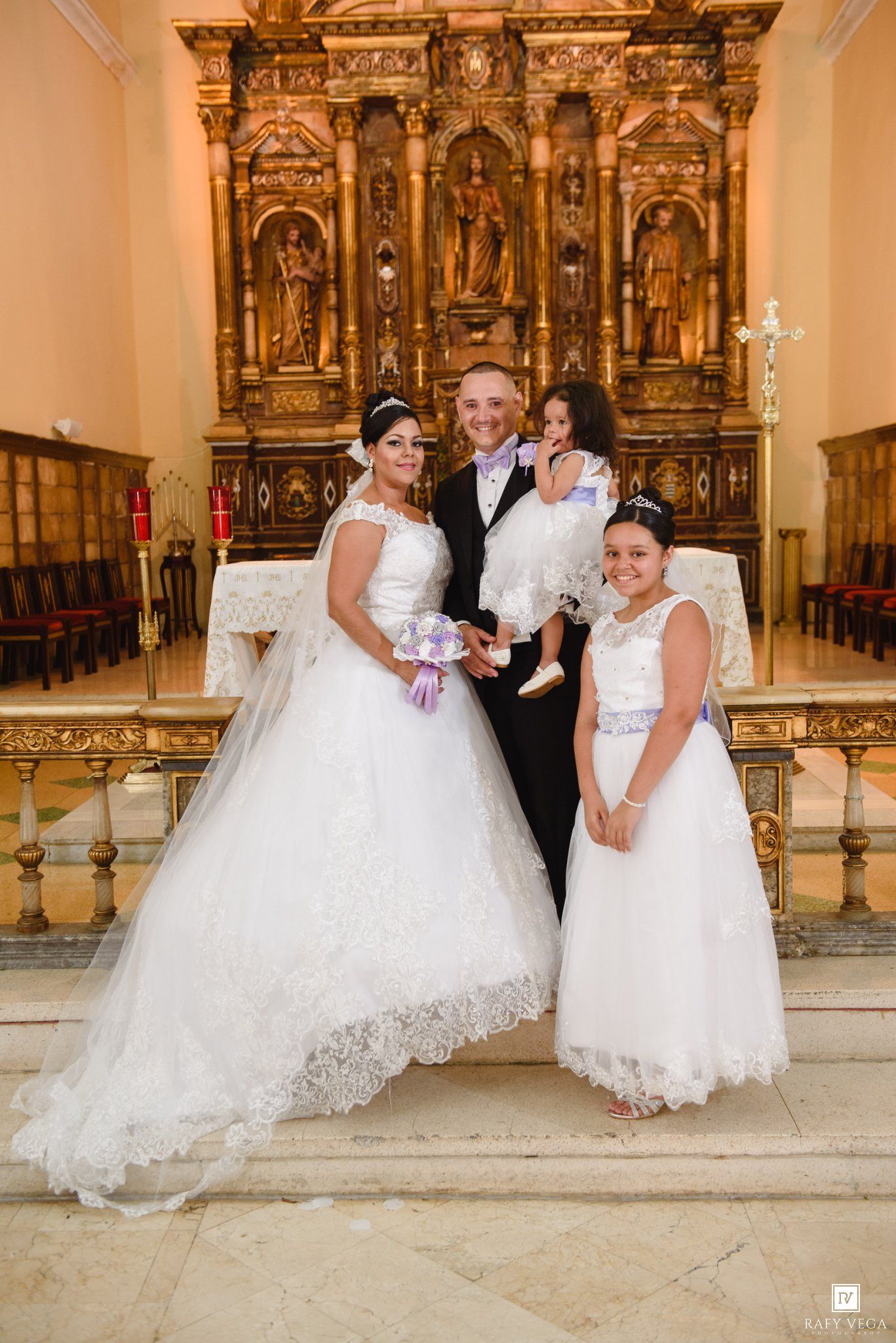 Wedding party poses before ornate golden altar, bride in ball gown, groom in suit, two girls in white dresses.