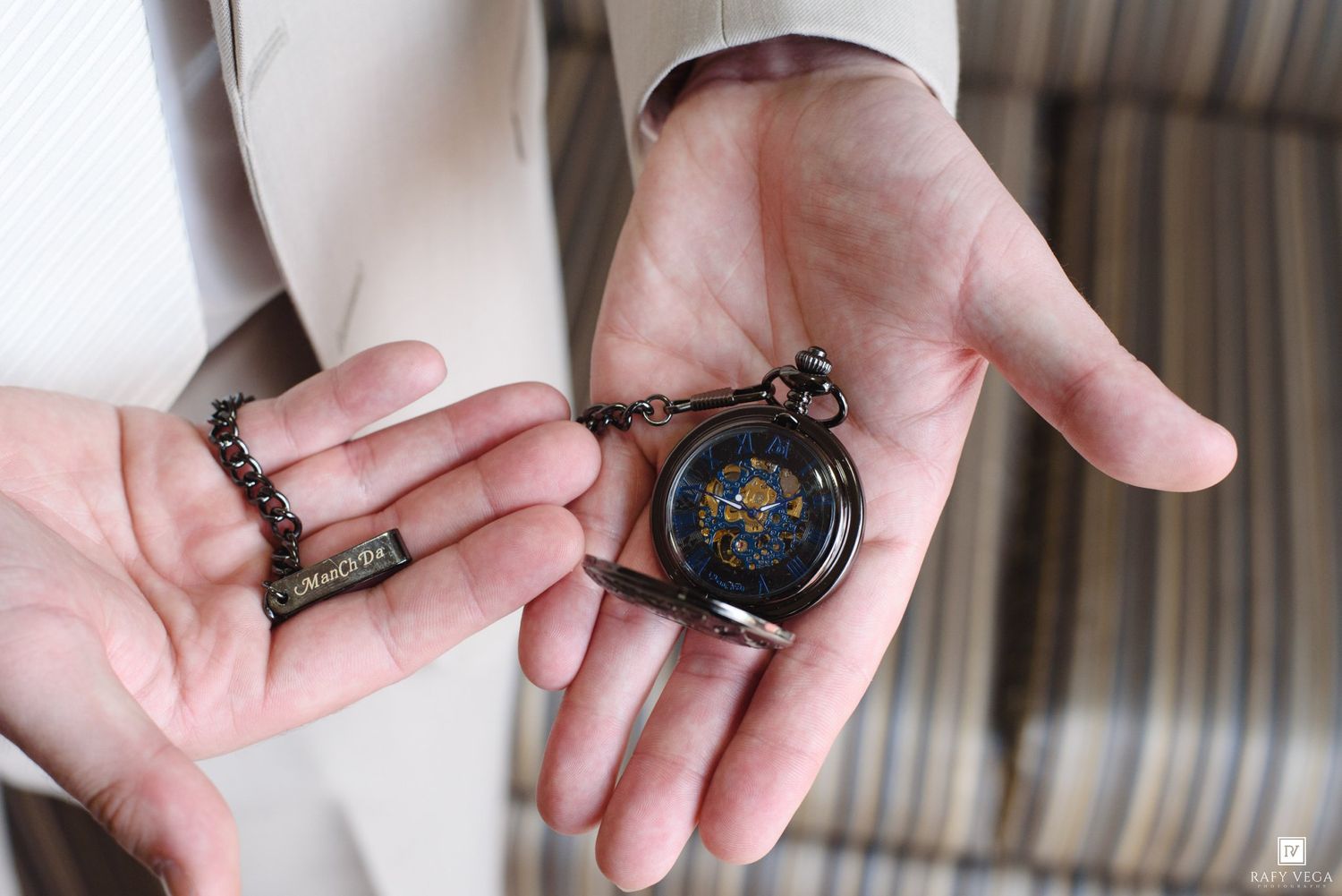 Groom holding a vintage black pocket watch with chain in open palms on wedding day.