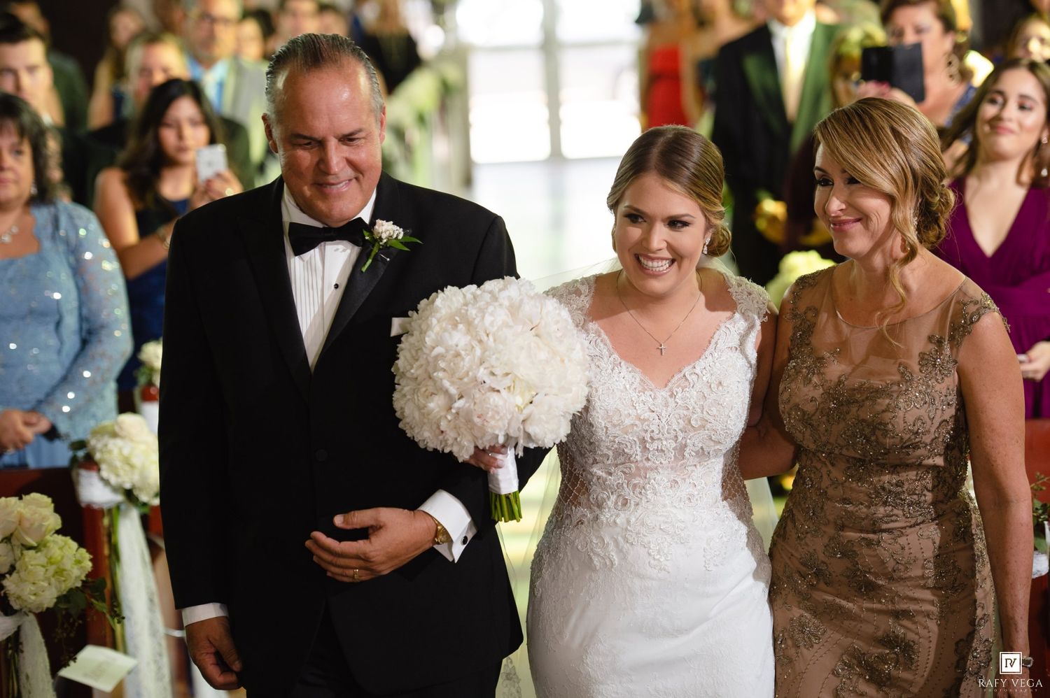 Bride in white lace gown walks down aisle with parents, holding white floral bouquet at elegant wedding ceremony.