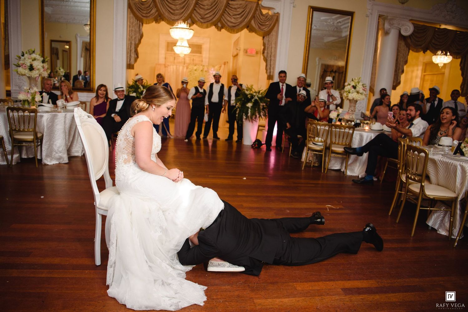Groom retrieves garter from bride sitting in chair at elegant wedding reception ballroom.