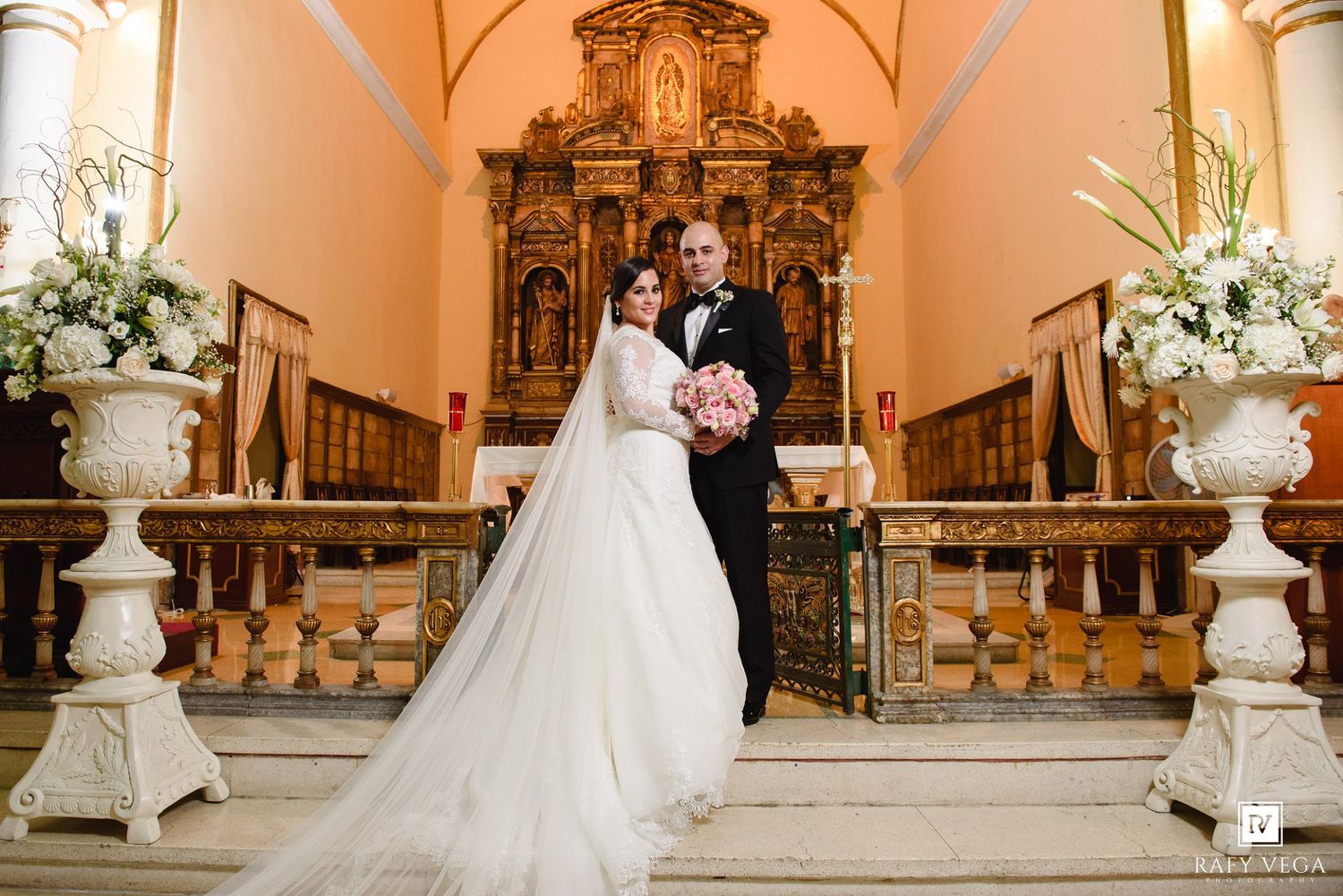 Wedding couple poses before golden baroque church altar flanked by white floral arrangements, bride in flowing gown.