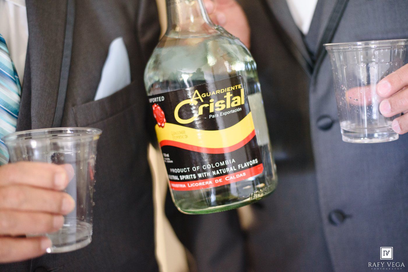 Two men in suits holding plastic cups and a bottle of Aguardiente Cristal Colombian spirit at a celebration.