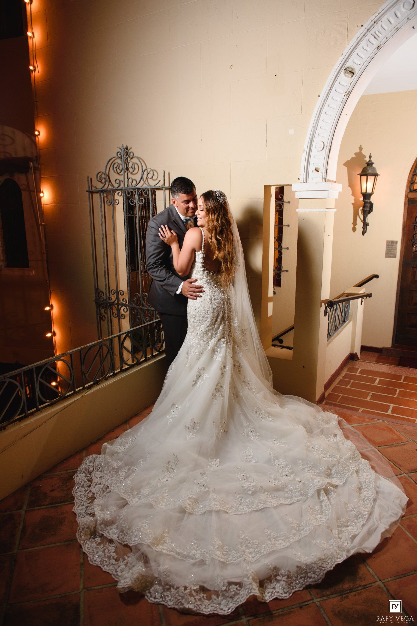 Bride in lace mermaid gown with cathedral veil embraces groom in gray suit near ornate arch in elegant venue.