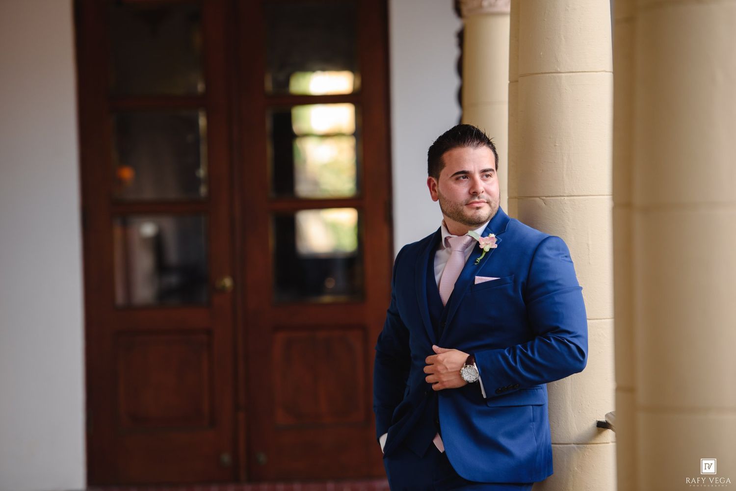 Groom in navy blue suit with pink tie and boutonniere standing by wooden doors.