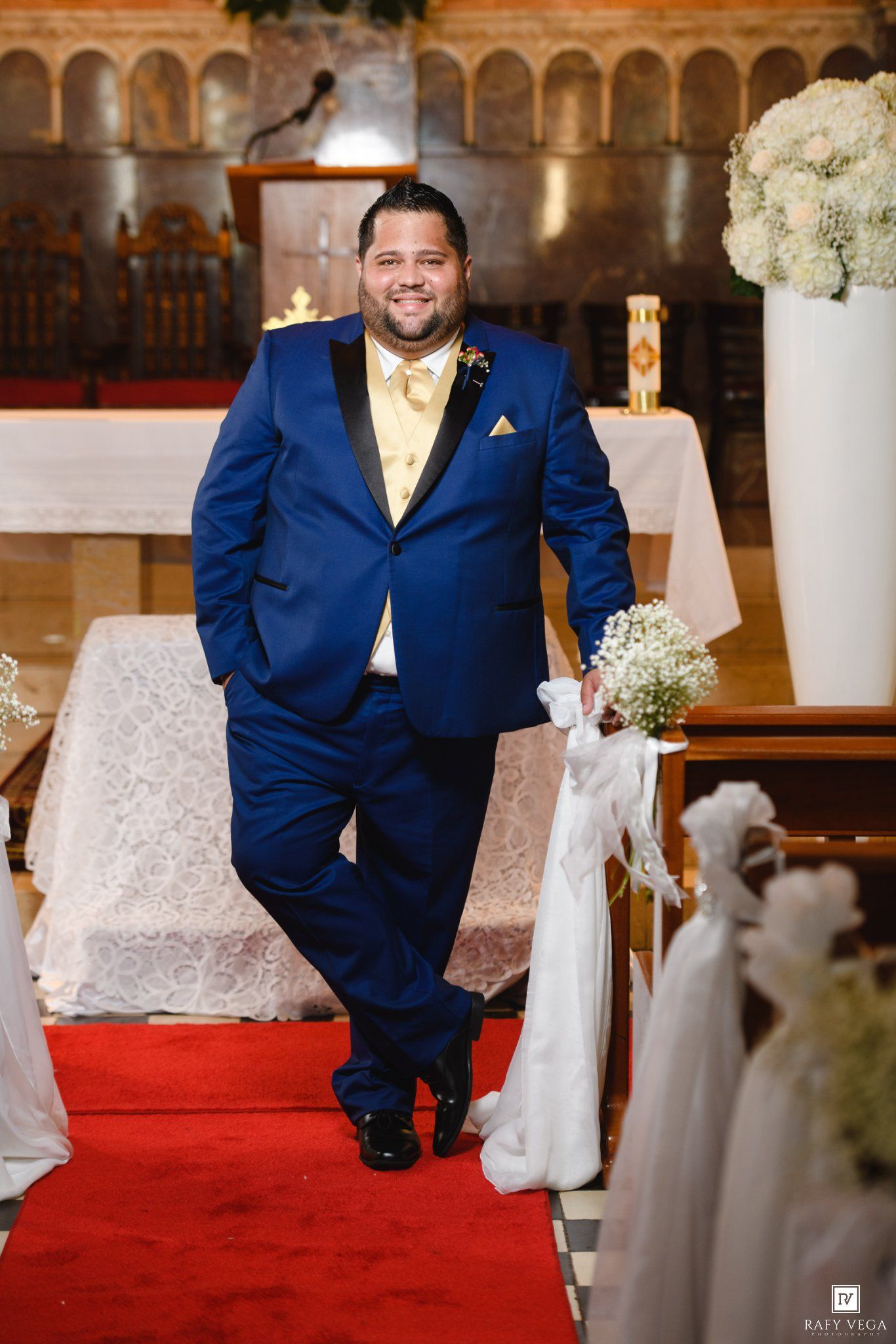 Groom in blue tuxedo with gold tie walks down red carpet aisle in decorated church on wedding day.