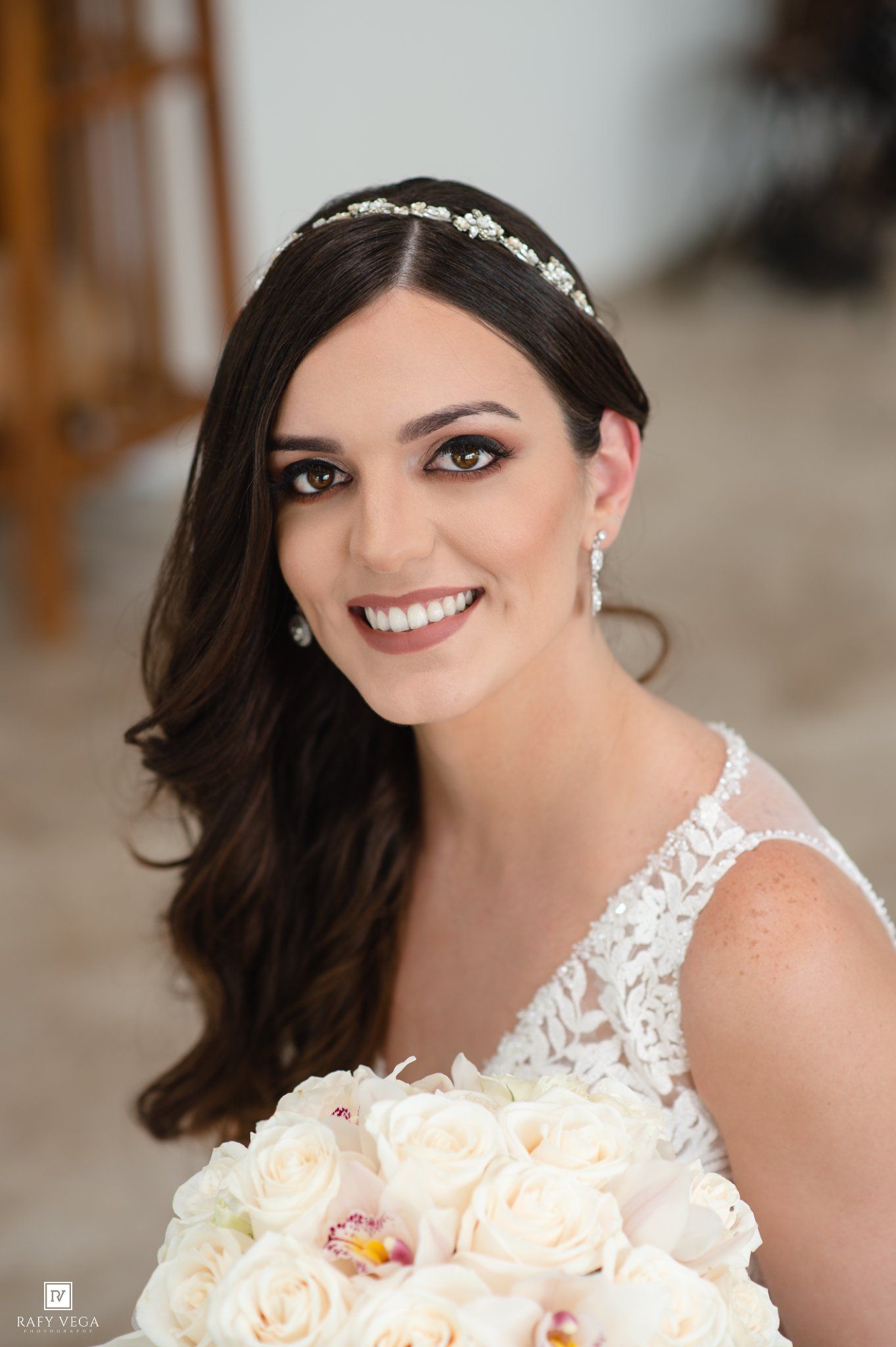 Smiling bride in lace dress holding white rose bouquet, wearing pearl earrings and floral headband.