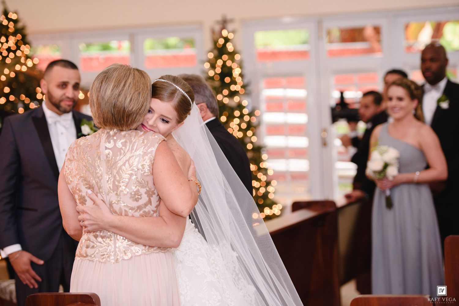 Bride in white gown and veil hugs a guest during wedding ceremony with bridesmaids and Christmas trees in background.