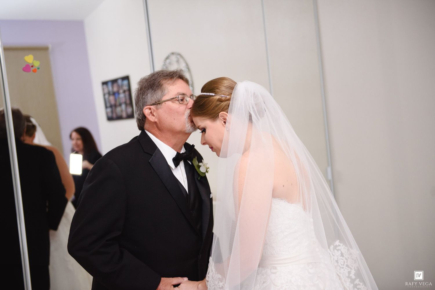 Father kisses bride on forehead in emotional wedding day moment, bride wearing white lace gown and veil.