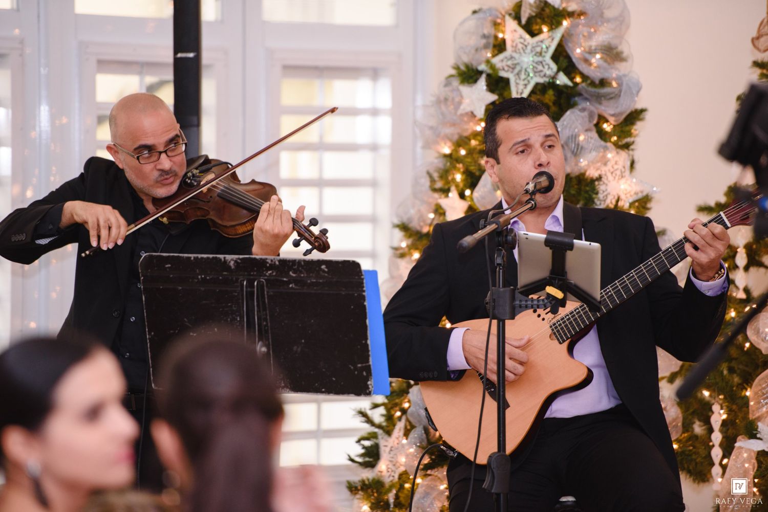 Two musicians perform holiday music, one playing violin and another singing with guitar near a decorated Christmas tree.