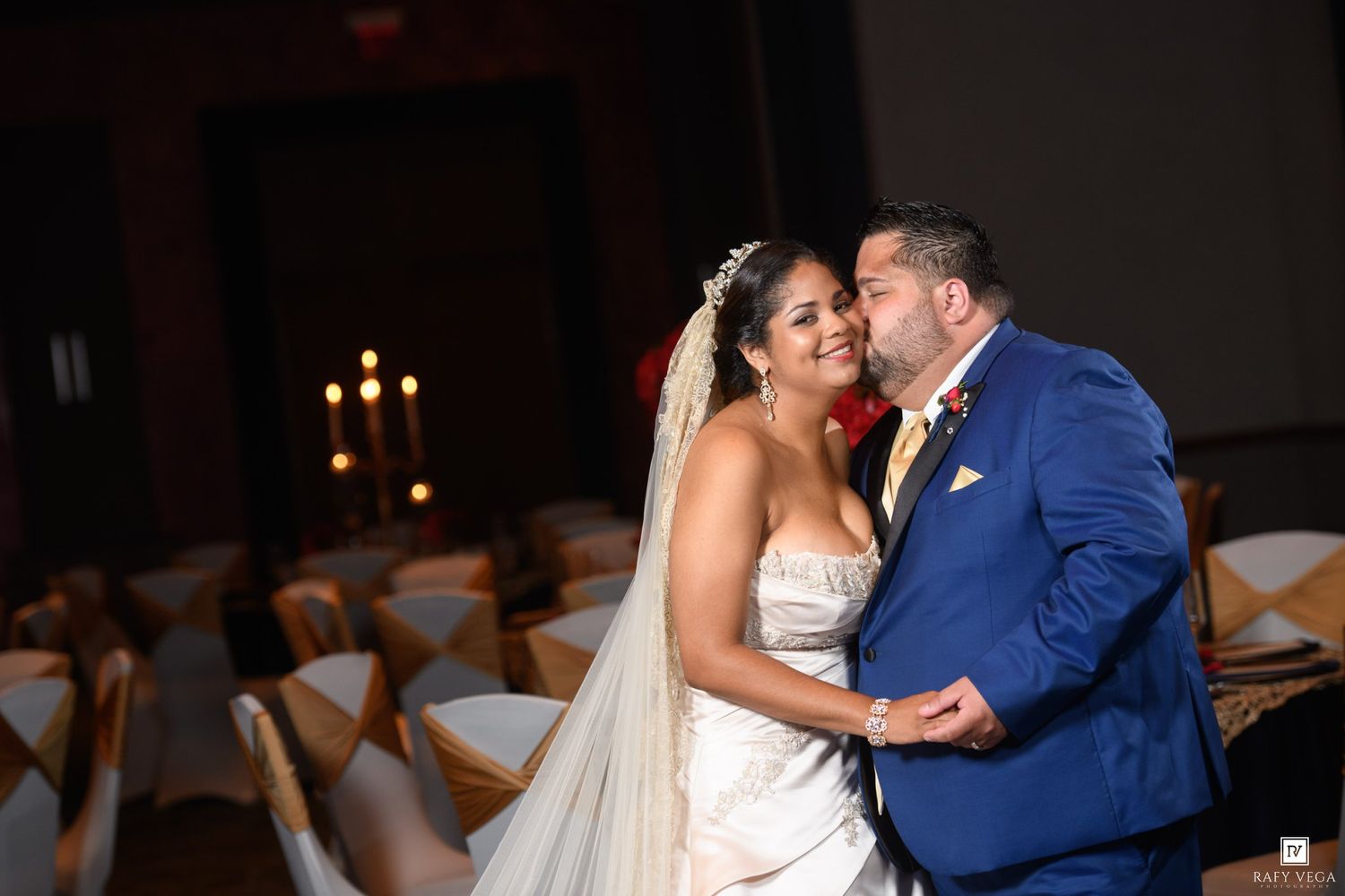 A bride and groom share a romantic moment in a decorated wedding reception hall, with the groom in a blue suit and bride in a white gown.