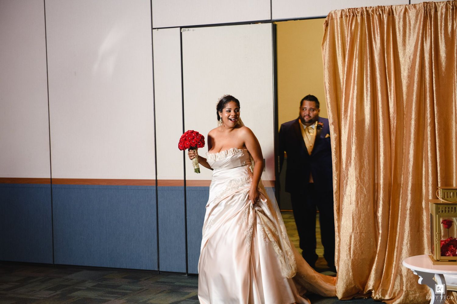 Bride in strapless gown holding red bouquet walks with groom in suit through gold curtained venue entrance.