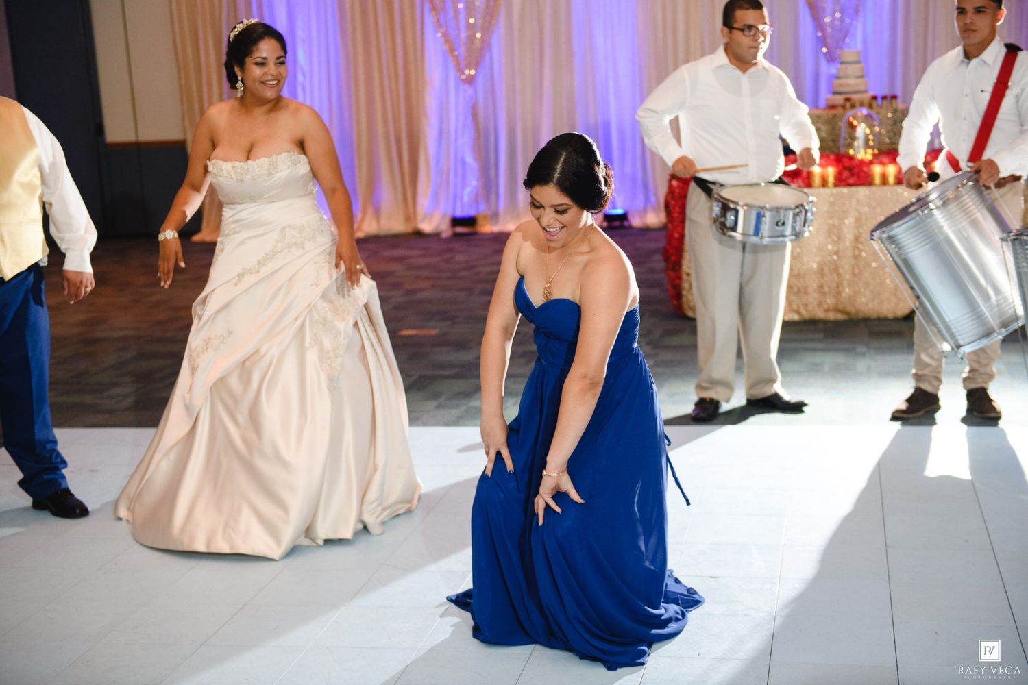 Bride and bridesmaid in blue gown dancing joyfully at wedding reception with drummer performing in background.