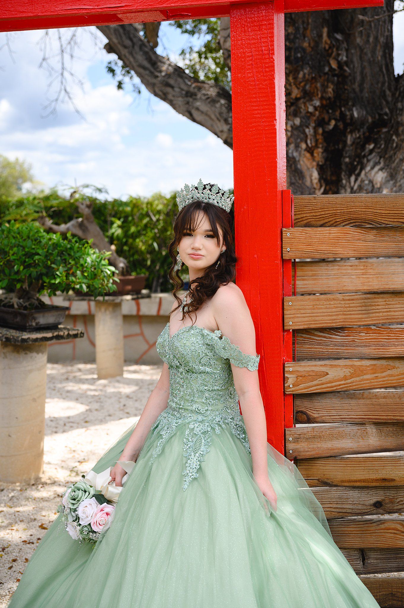 Young woman in sage green quinceañera ball gown with crown and pink bouquet poses by red wooden structure.