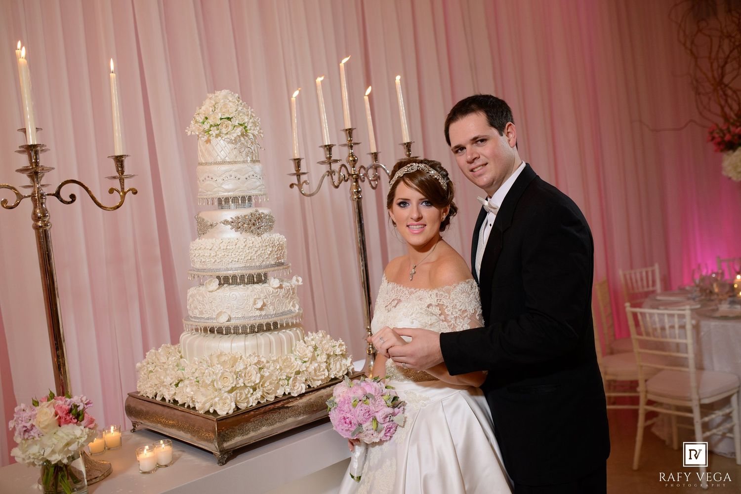 Bride and groom smiling beside a tall elegant white wedding cake with candelabras in a pink draped reception hall.