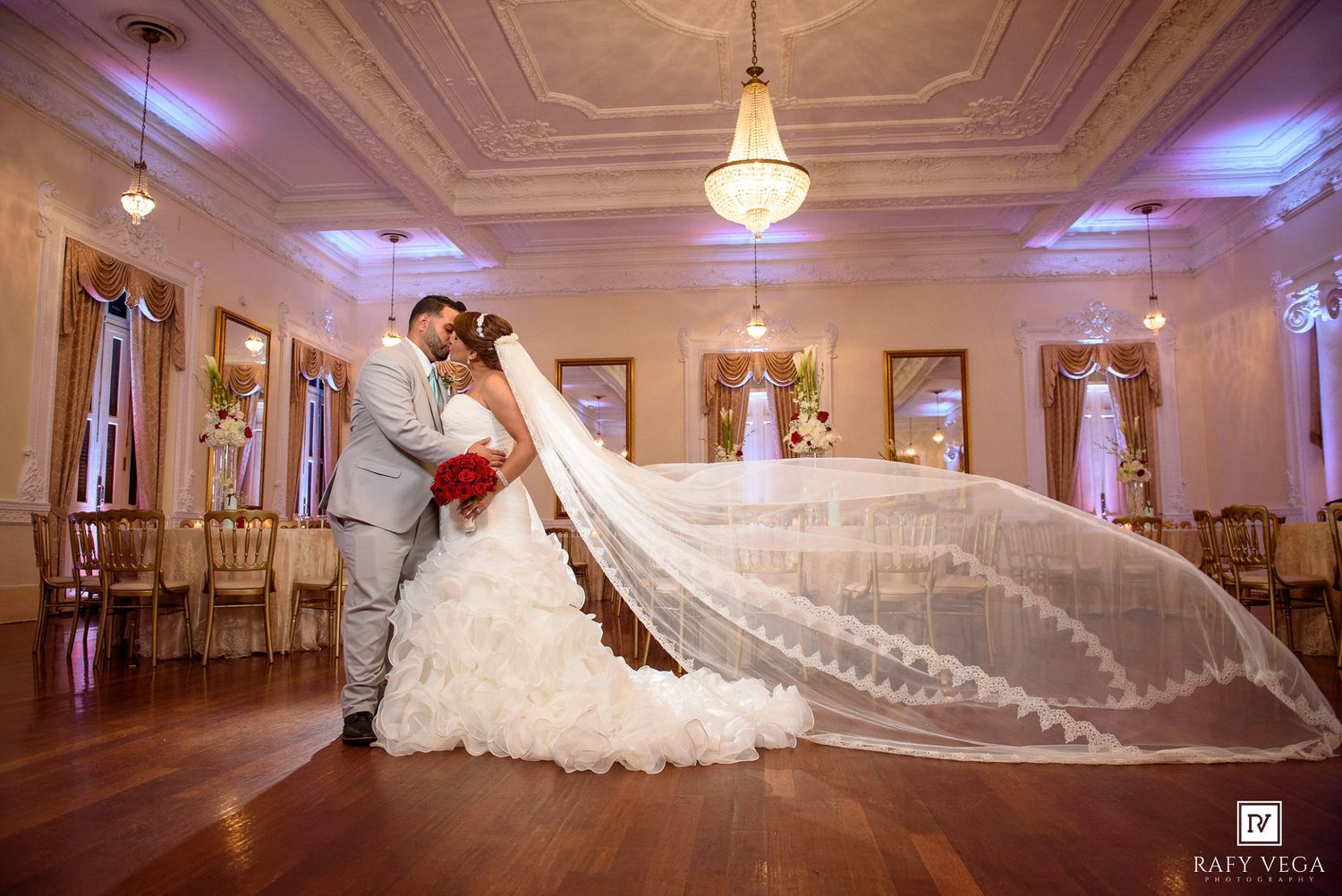 Bride and groom share romantic first dance kiss, cathedral veil flowing dramatically in elegant ballroom wedding venue.