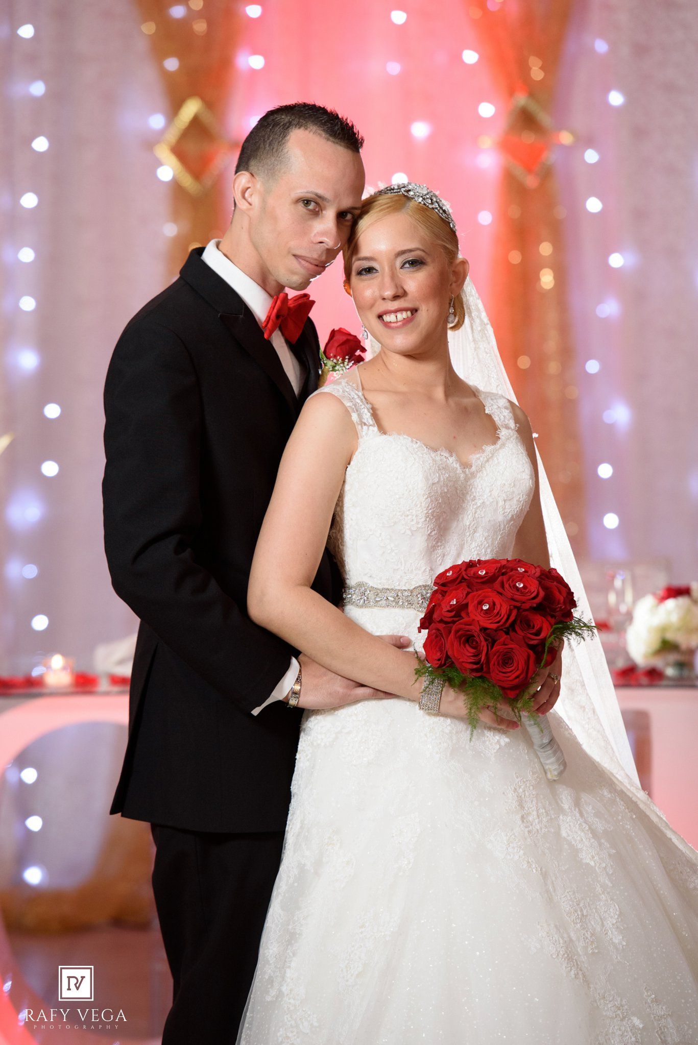 Bride in white gown holding red rose bouquet poses with groom in black tuxedo at elegant wedding reception.