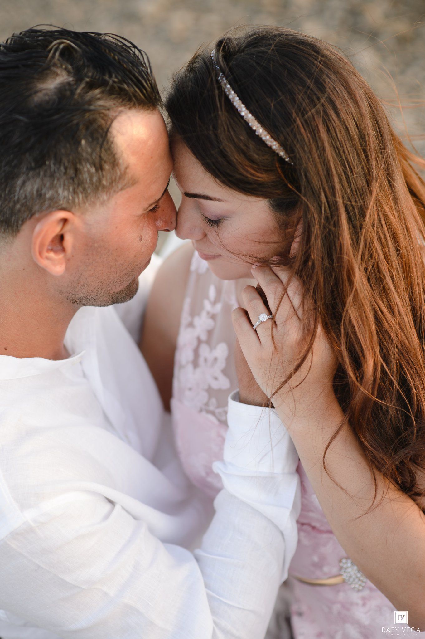 Romantic couple in white attire touching foreheads tenderly, woman wearing engagement ring and headband outdoors.