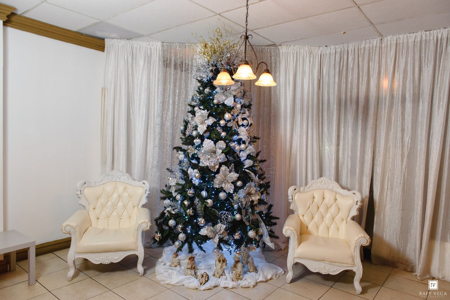Elegant Christmas tree with white and gold decorations flanked by two white tufted chairs in a festive holiday setting.