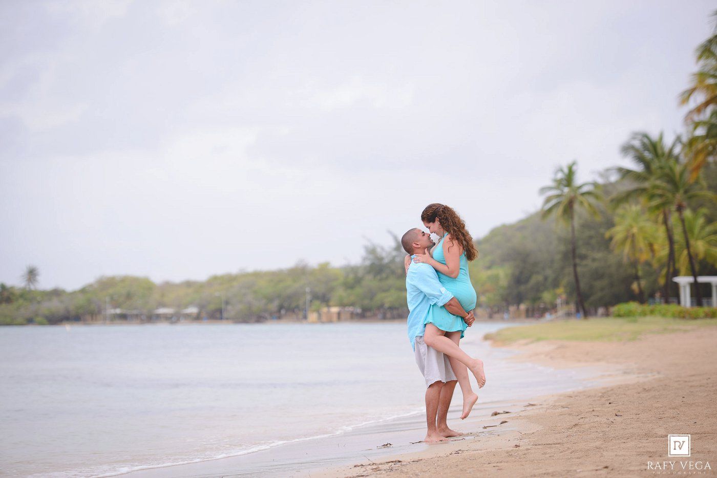 Couple embracing on a tropical beach, man lifting woman in matching turquoise outfits with palm trees and ocean backdrop.