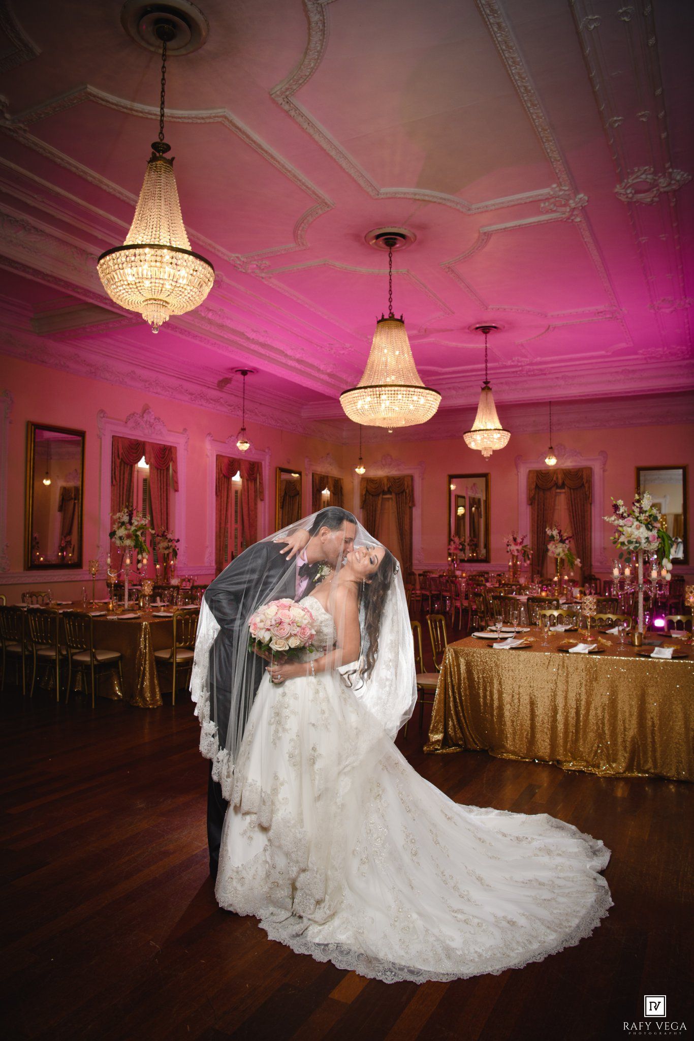 Bride and groom share romantic kiss in elegant ballroom with crystal chandeliers, pink uplighting, and gold sequin tablecloths.