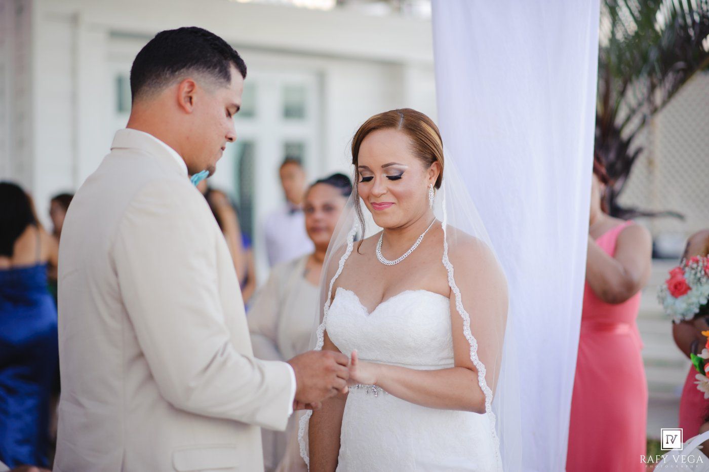 A bride and groom exchange rings during an outdoor wedding ceremony, with guests visible in the background.