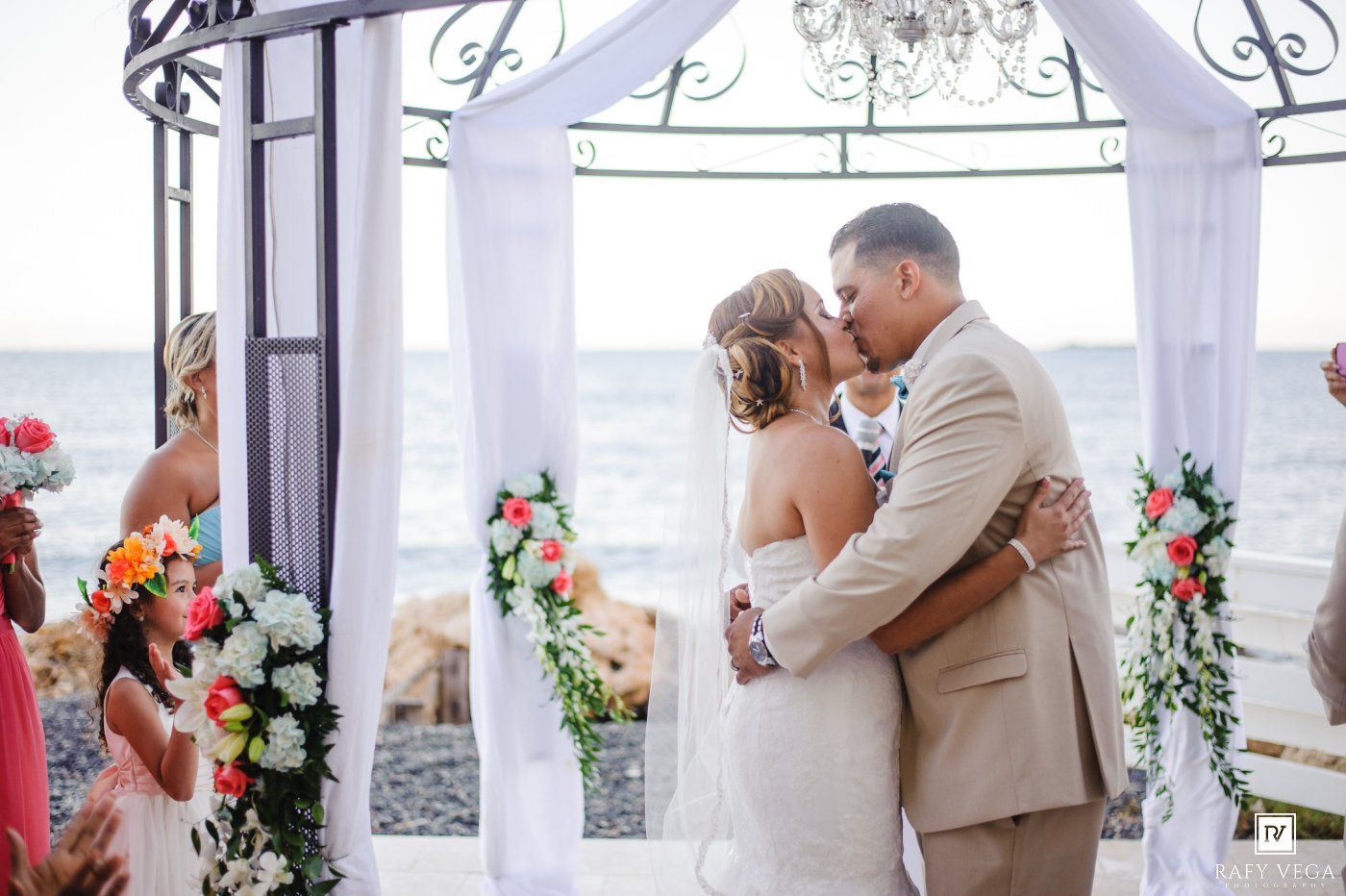 Bride and groom share first kiss at beachside wedding ceremony under white draped gazebo with colorful floral arrangements.