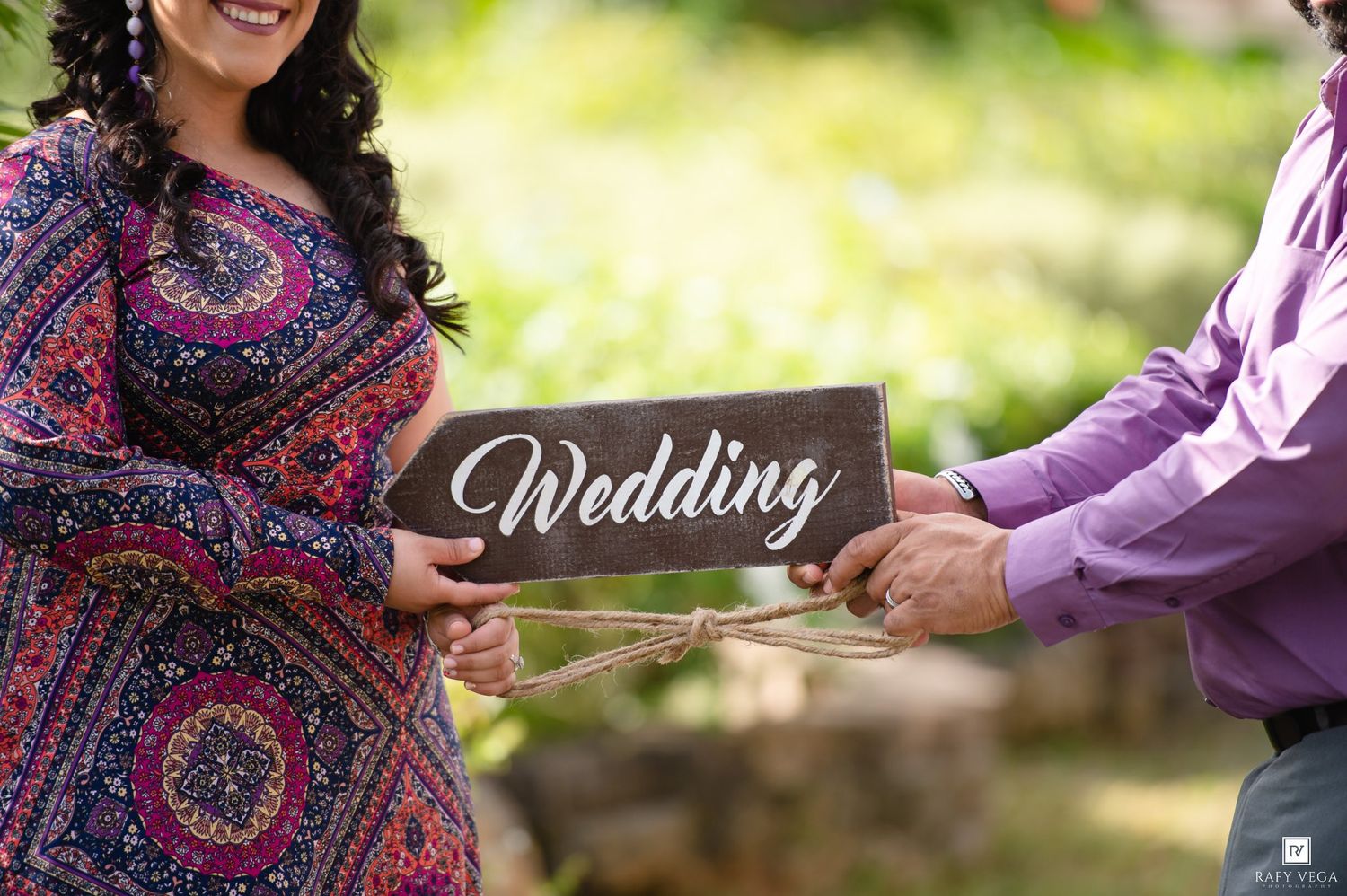 Couple holding a rustic wooden Wedding sign outdoors, woman in colorful dress and man in purple shirt.