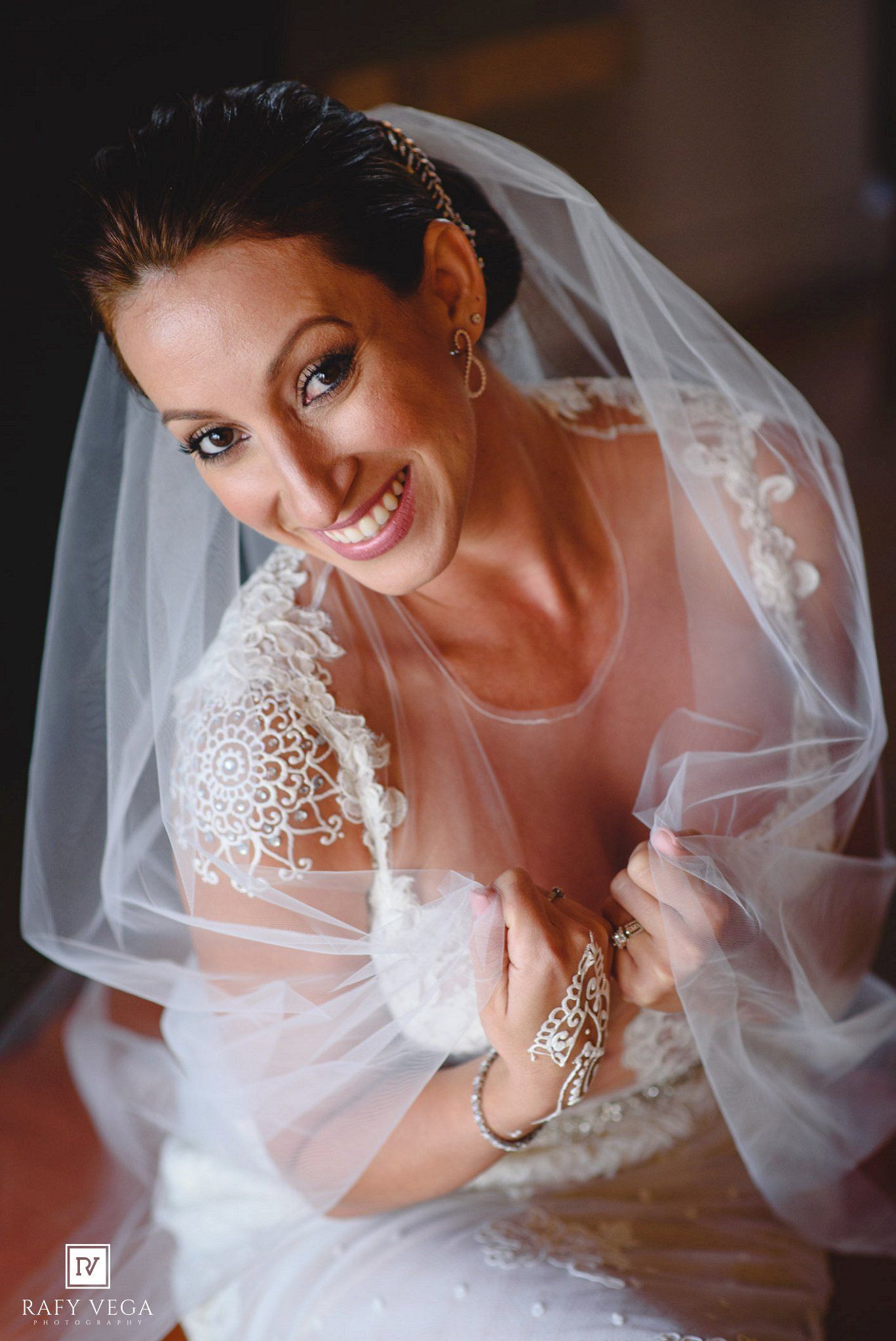 Smiling bride in lace wedding dress and veil poses elegantly, showcasing bridal jewelry and makeup on her wedding day.