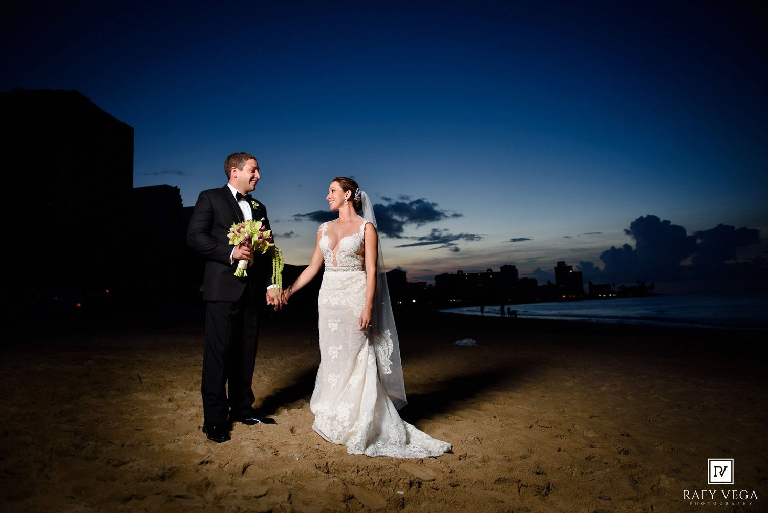 Bride and groom hold hands on a sandy beach at twilight, with a glowing sunset sky behind them.