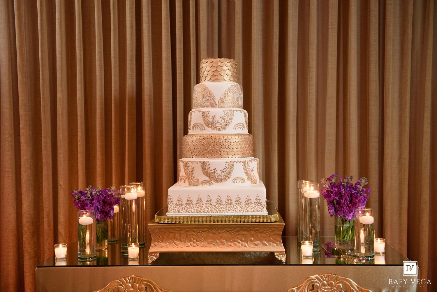 Elegant six-tier white and gold wedding cake on a mirrored table flanked by purple flowers and candles against draped curtains.