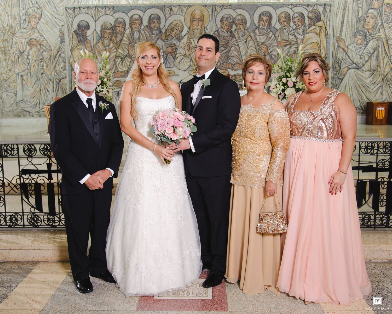 Wedding party of five posing in church, bride in white gown holding pink bouquet, groom in tuxedo, with family members.