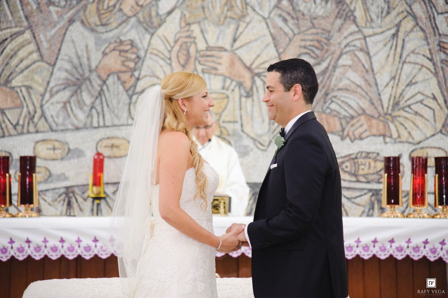 Bride and groom exchange vows at church altar with mosaic mural backdrop during wedding ceremony.