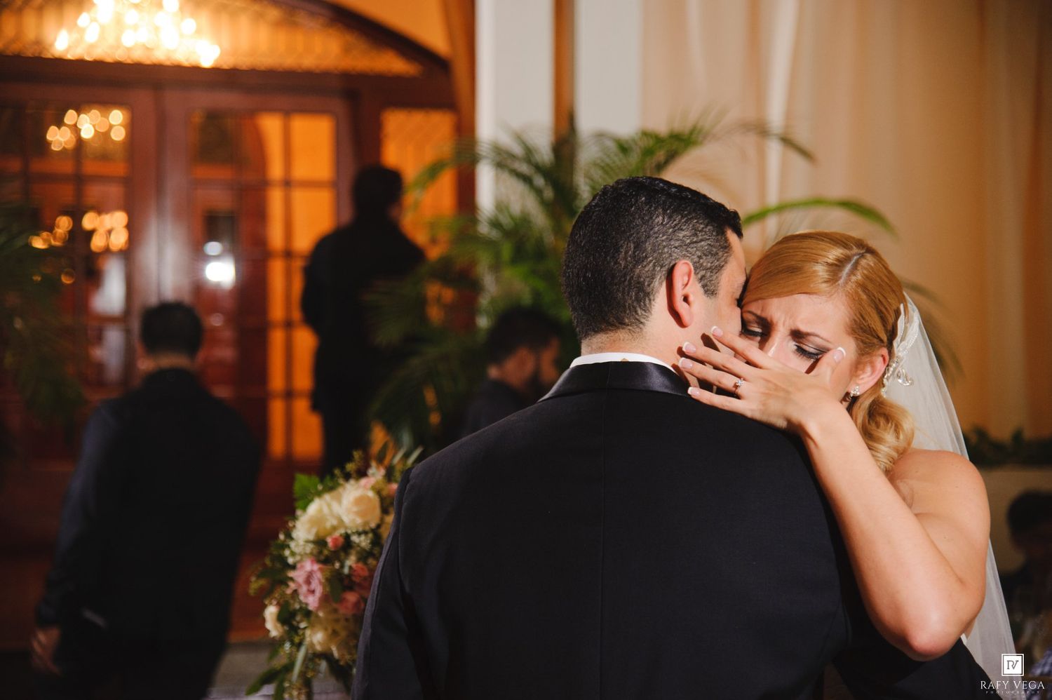 Bride and groom share emotional first dance embrace at elegant wedding reception with floral arrangements.