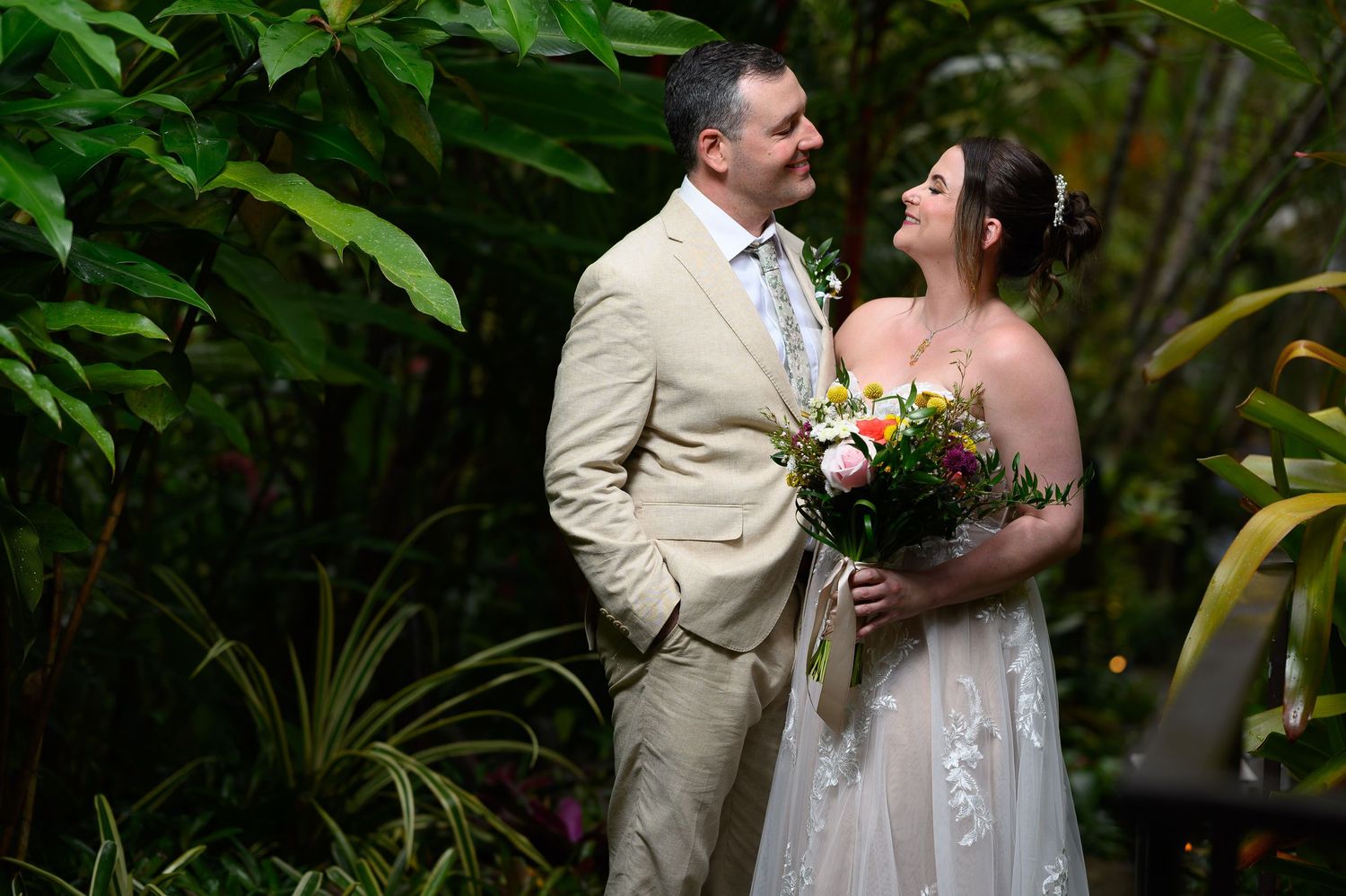 Bride and groom share a romantic moment surrounded by lush tropical greenery, bride holding a colorful bouquet.