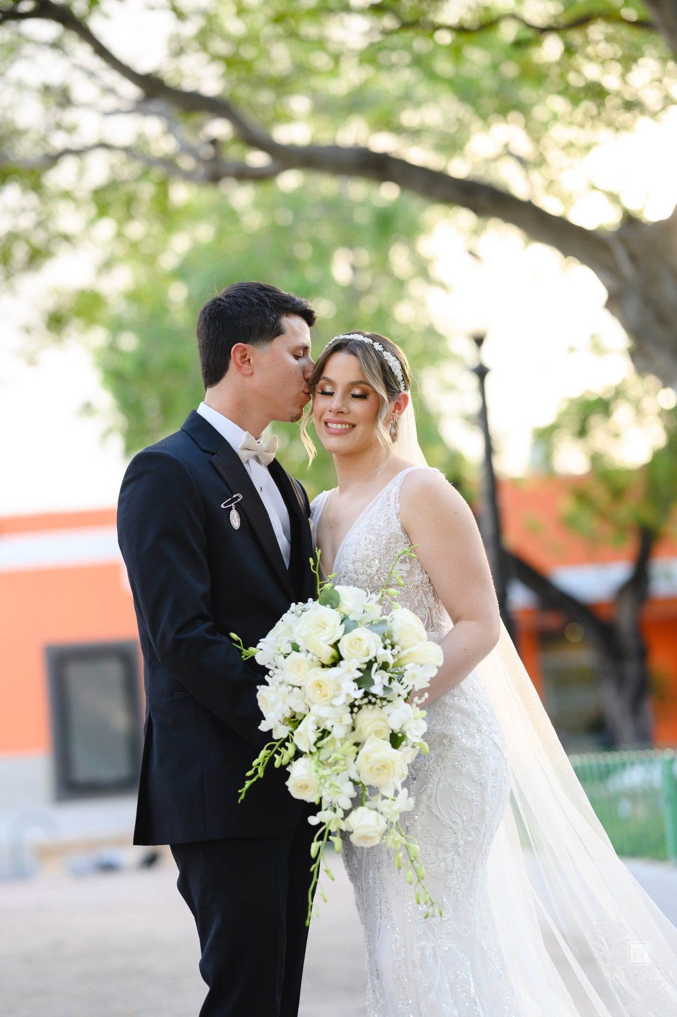 Bride in lace gown holds white bouquet as groom in black tuxedo kisses her cheek outdoors.