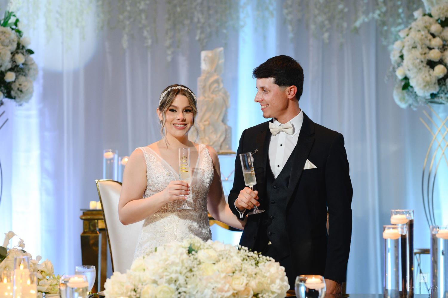 Bride and groom toasting with champagne at luxurious white floral wedding reception table.