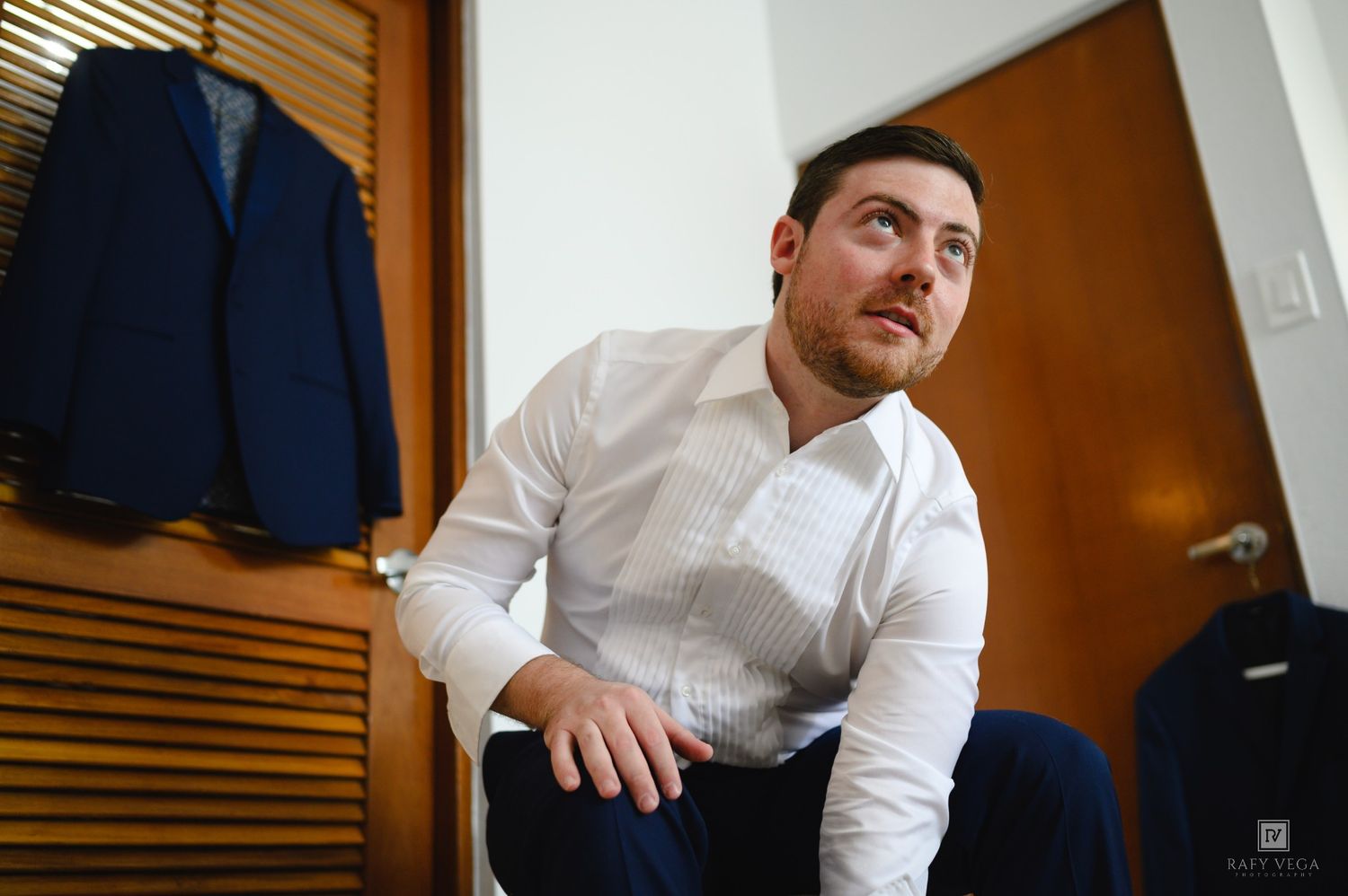 Man in white dress shirt sitting casually near a blue suit jacket hanging on a wooden wardrobe.
