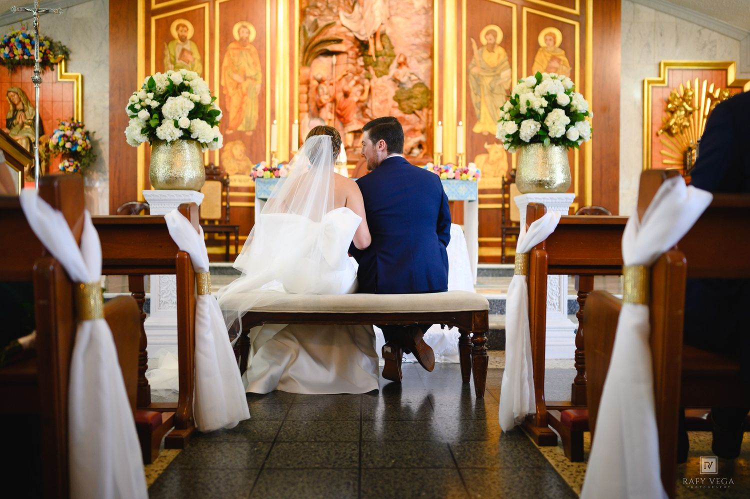 Bride and groom sitting together at church altar during wedding ceremony with white floral arrangements.