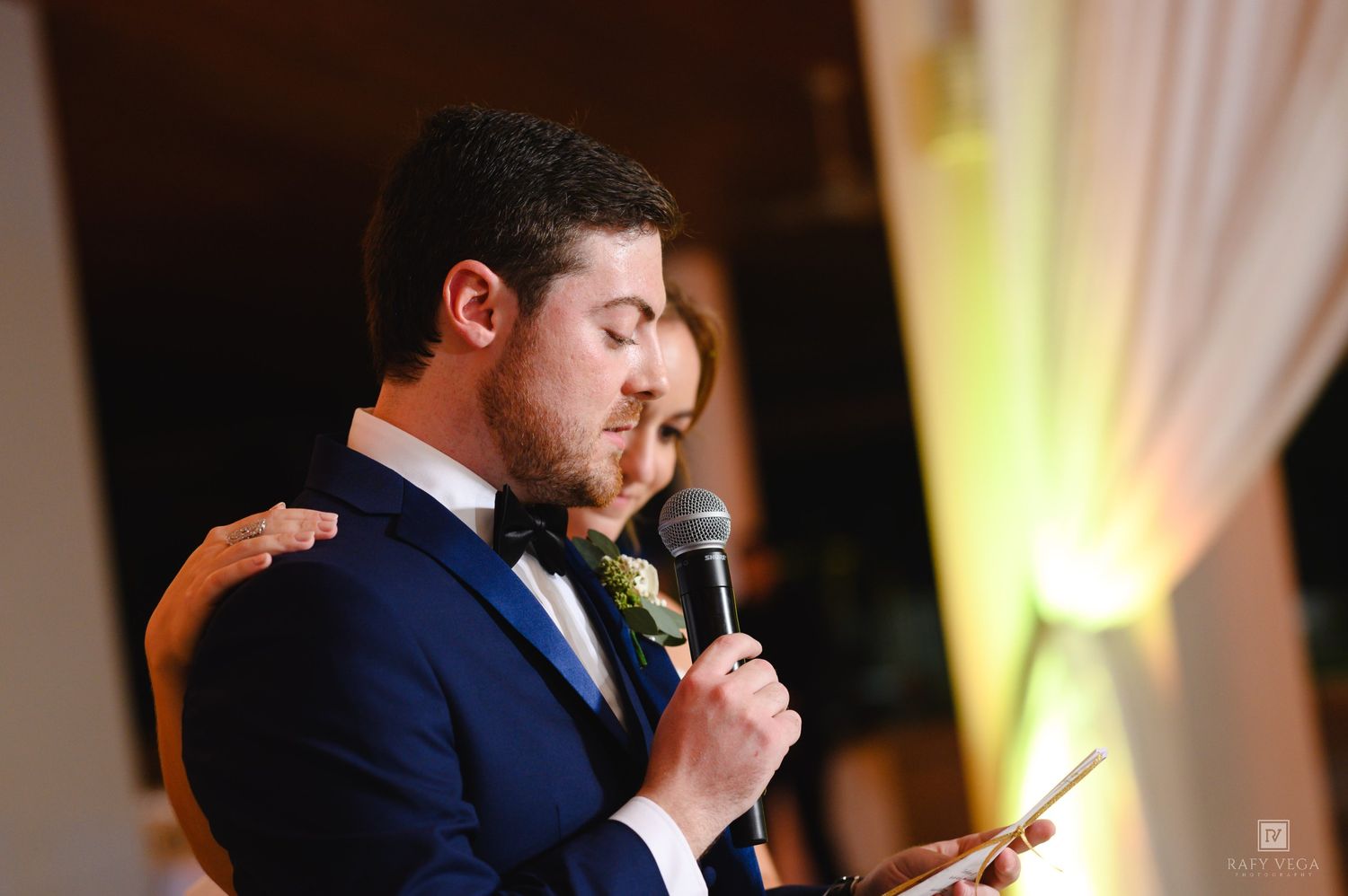 Groom in navy suit reads heartfelt vows into a microphone during wedding reception speech.