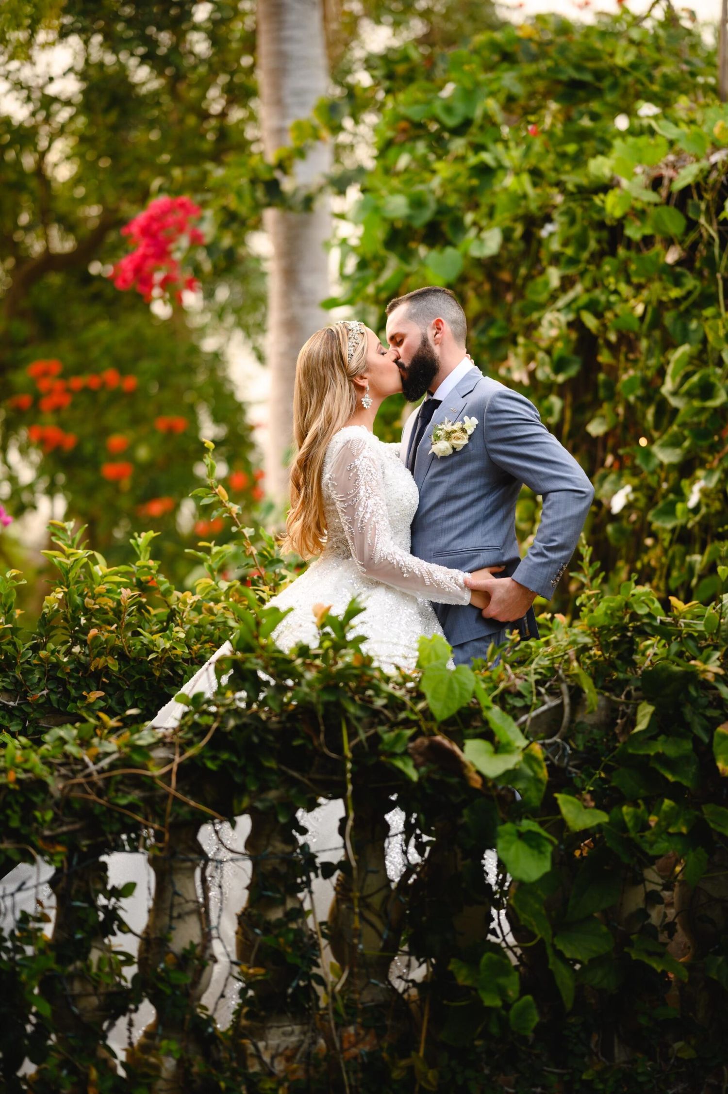 Bride and groom share a romantic kiss surrounded by lush green foliage and colorful flowers in a garden setting.