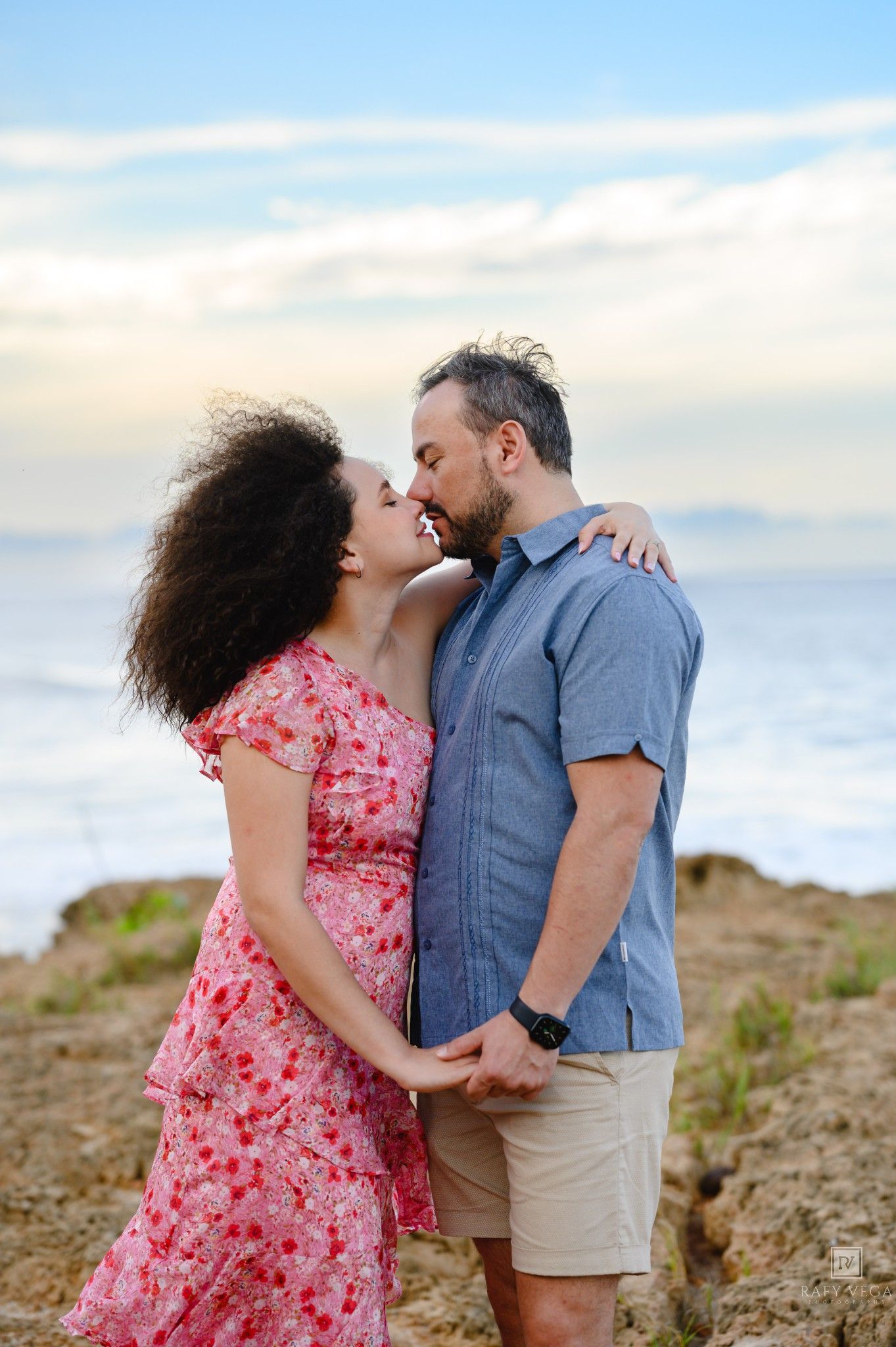 Romantic couple embracing and almost kissing on rocky beach at sunset, woman in floral dress, man in blue shirt.