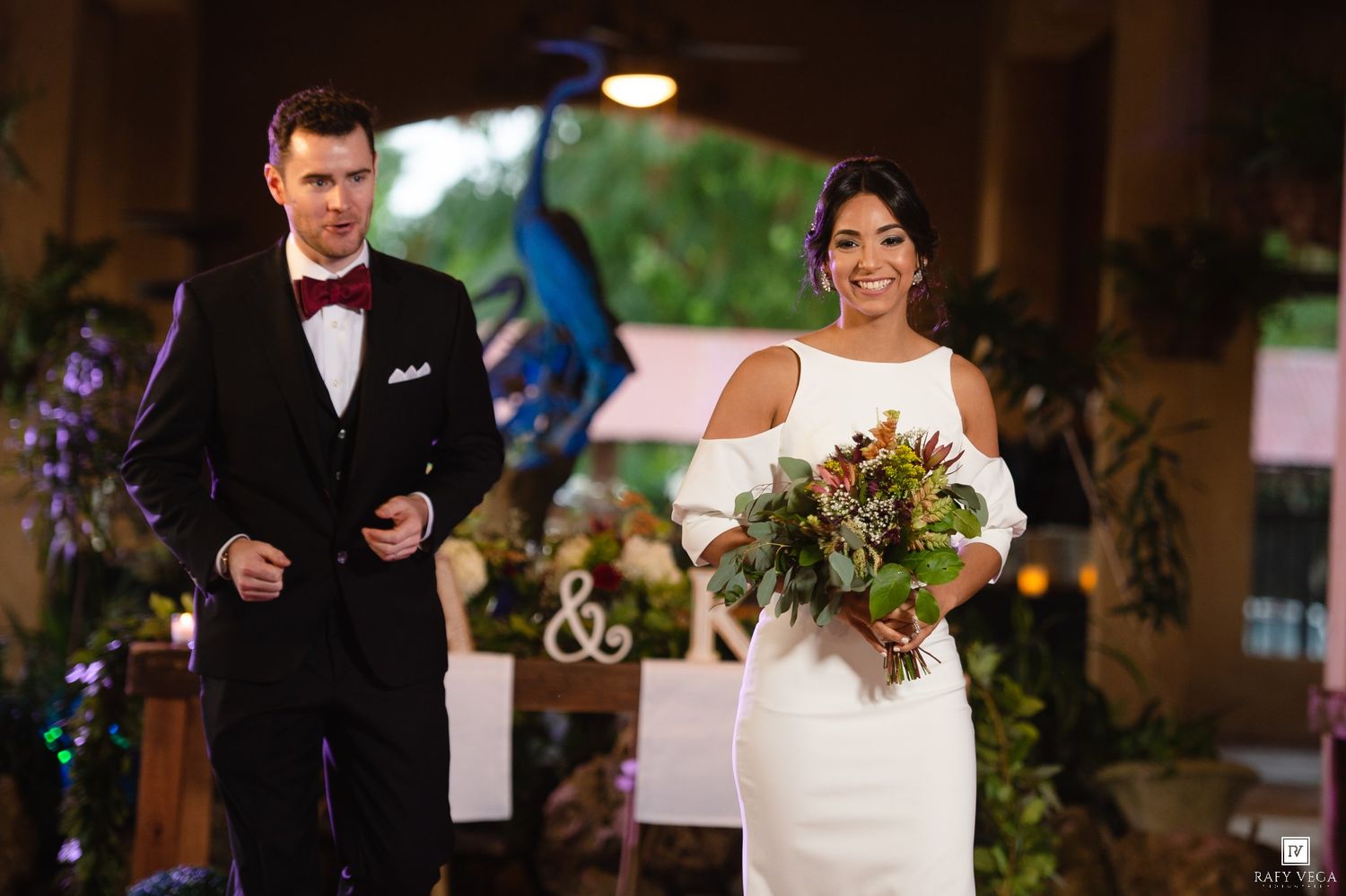 Bride in white off-shoulder gown holding bouquet walks with groom in black tuxedo at elegant wedding ceremony.
