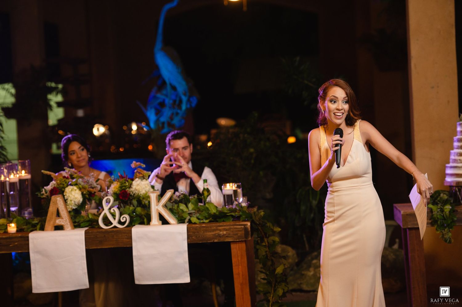 Woman in elegant gown gives a speech at a wedding reception while couple sits at decorated head table.