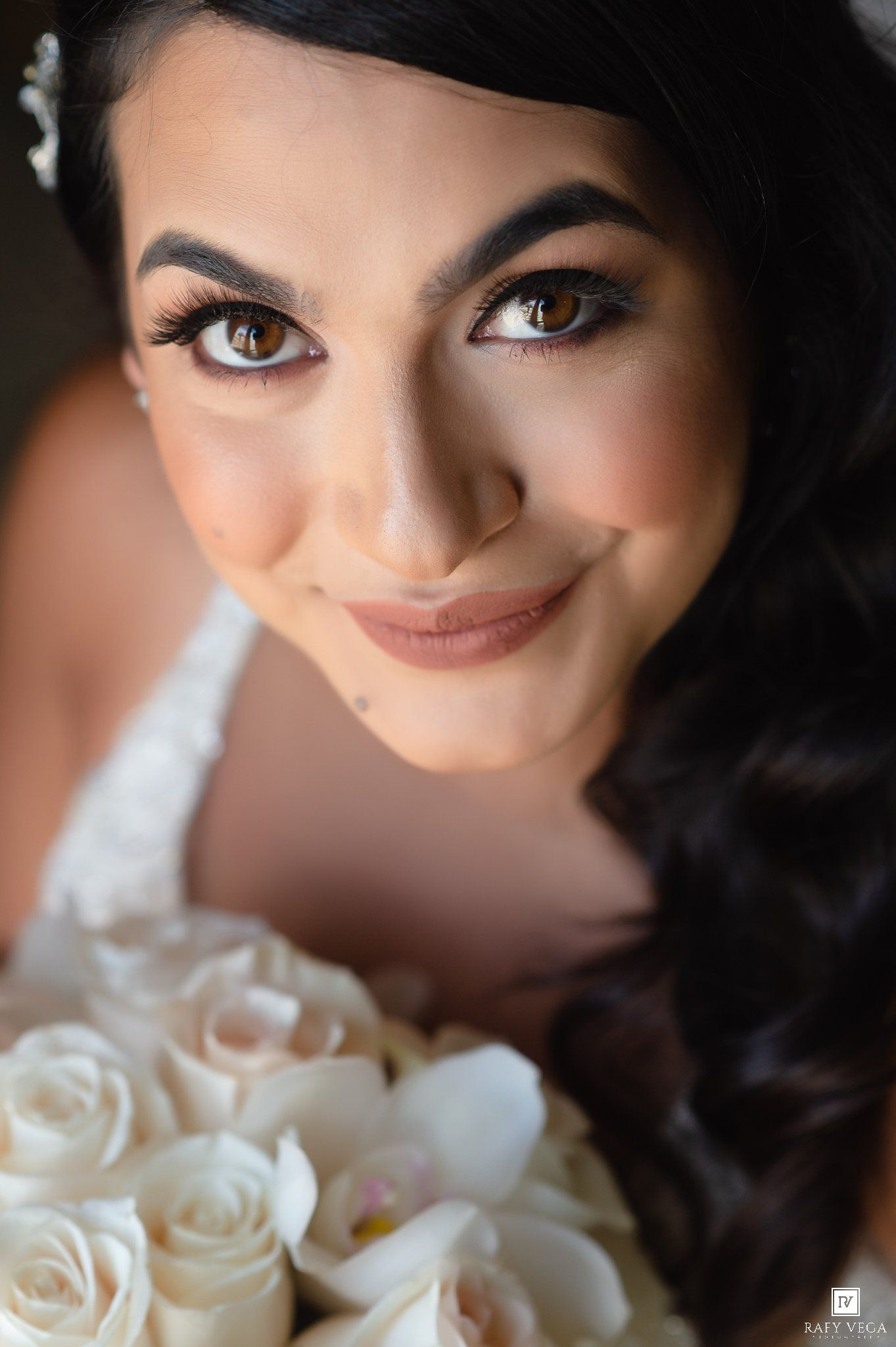 Close-up portrait of a smiling bride with dark hair and brown eyes holding a bouquet of white roses on her wedding day.